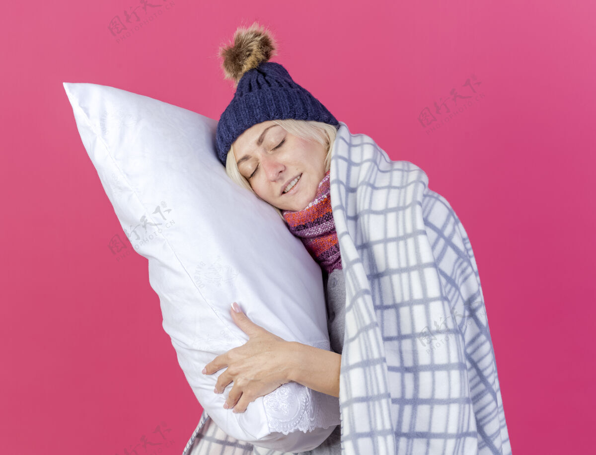 人年轻的金发病女人戴着冬天的帽子 围巾裹着格子布 高兴地把头放在枕头上 隔离在粉红色的墙上姿势金发冬天