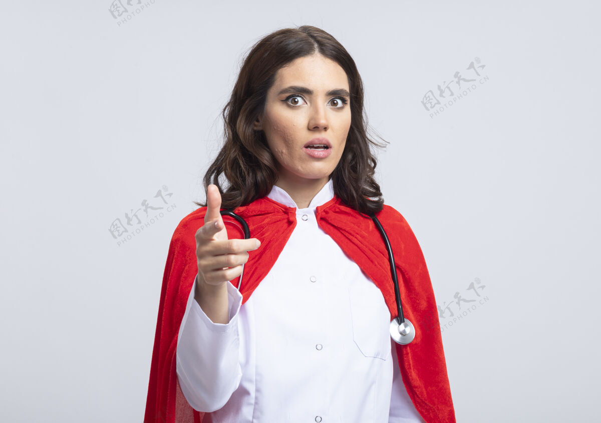 女孩焦虑的女超人穿着医生制服 头戴红斗篷 听诊器指向隔离在白墙上的前方姿势医生人