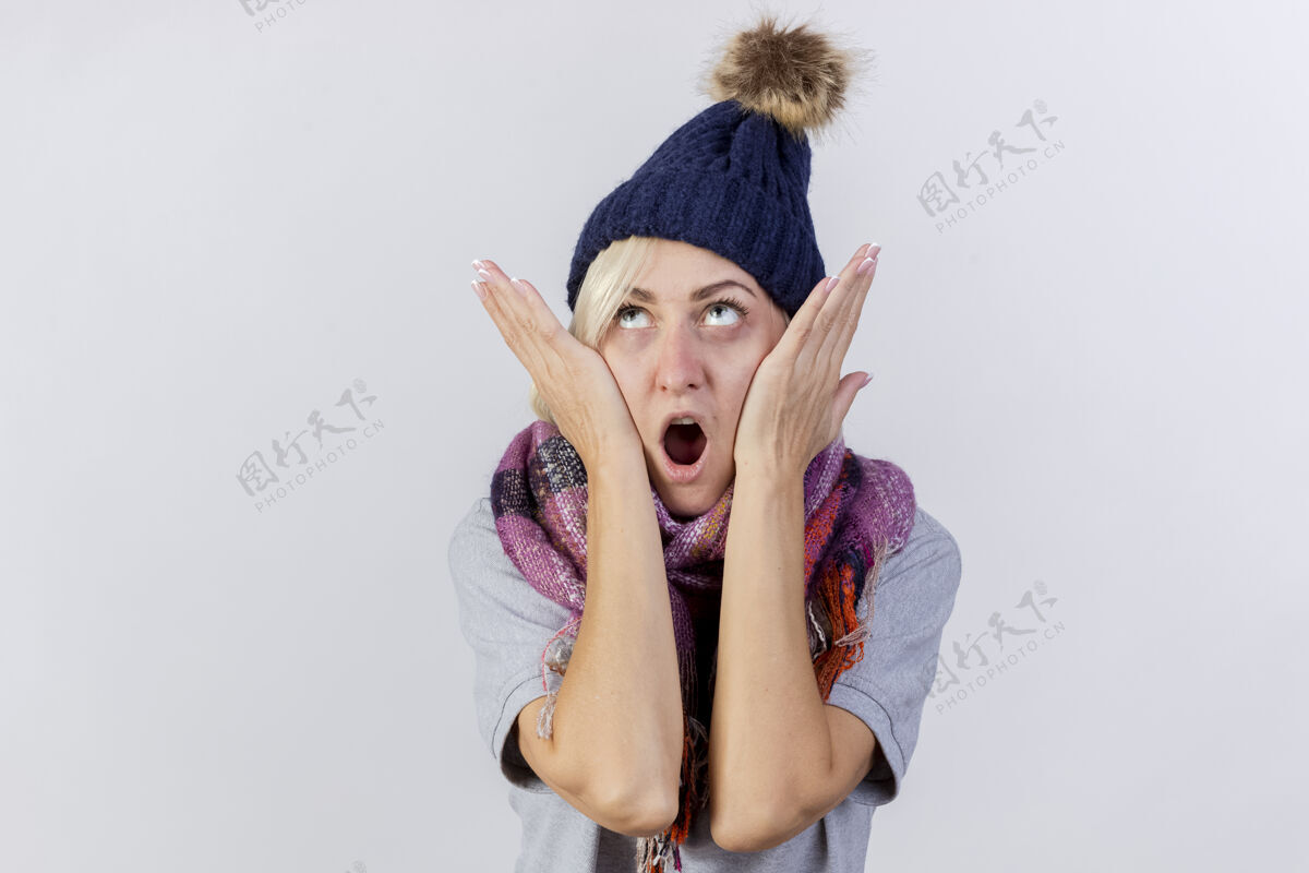 疾病震惊的年轻金发病女人戴着冬天的帽子和围巾把手放在脸上看着隔离的白色墙壁人帽子人