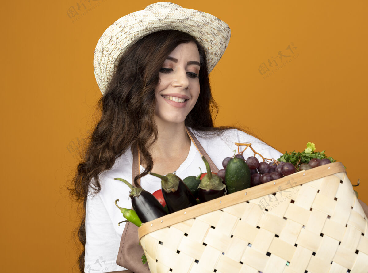 人年轻的女园丁面带微笑 穿着制服 戴着园艺帽 手里拿着 看着隔离在橙色墙上的菜篮子人花园穿着