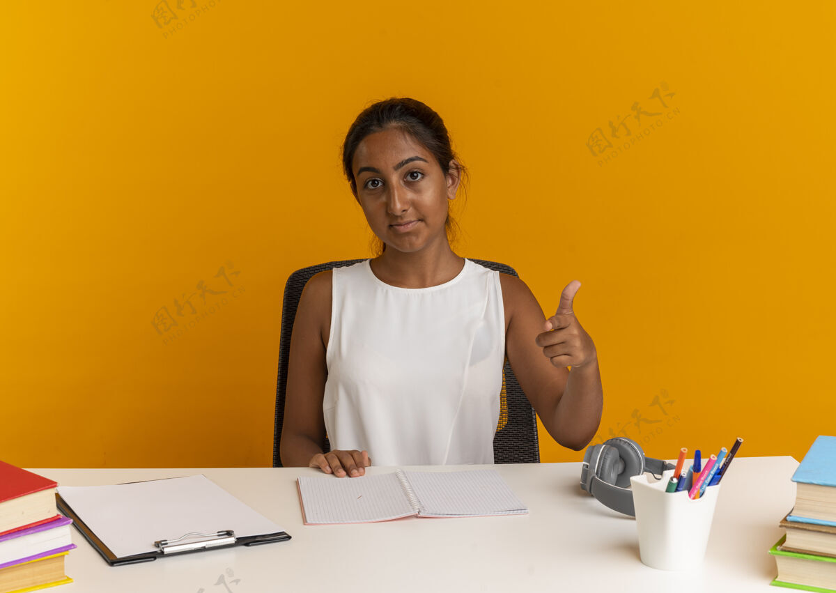 女学生看着镜头 坐在书桌旁的年轻女学生拿着学习工具向你展示姿势橙色学校手势