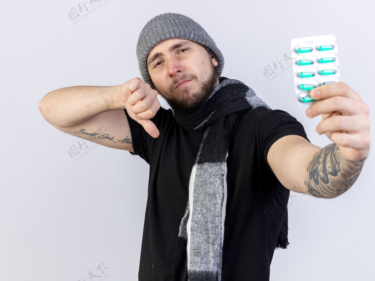 人戴着冬天的帽子和围巾的自信的年轻病人大拇指朝下 拿着隔离在白墙上的医药胶囊包穿人人