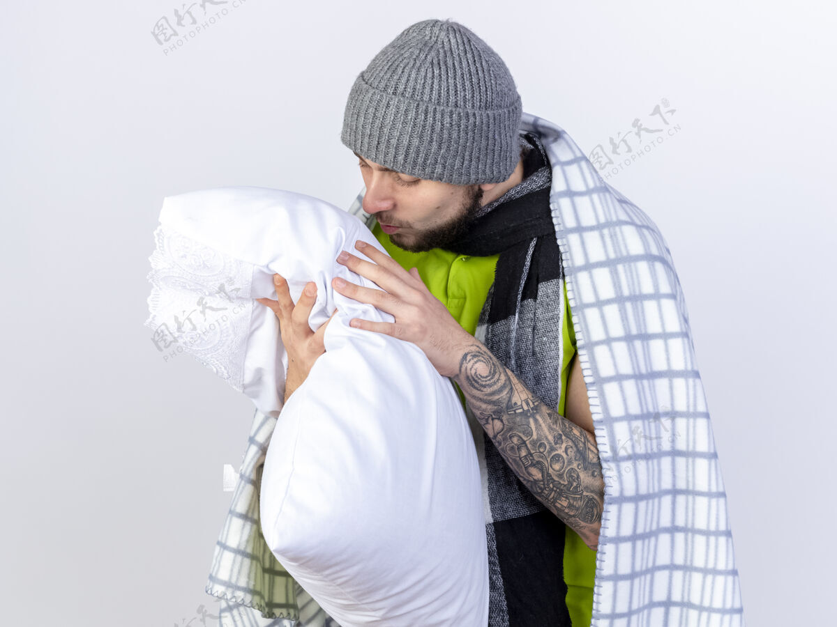姿势高兴的年轻人戴着冬天用格子布包裹的帽子抱着病号 看着隔离在白墙上的枕头年轻人脸拿着