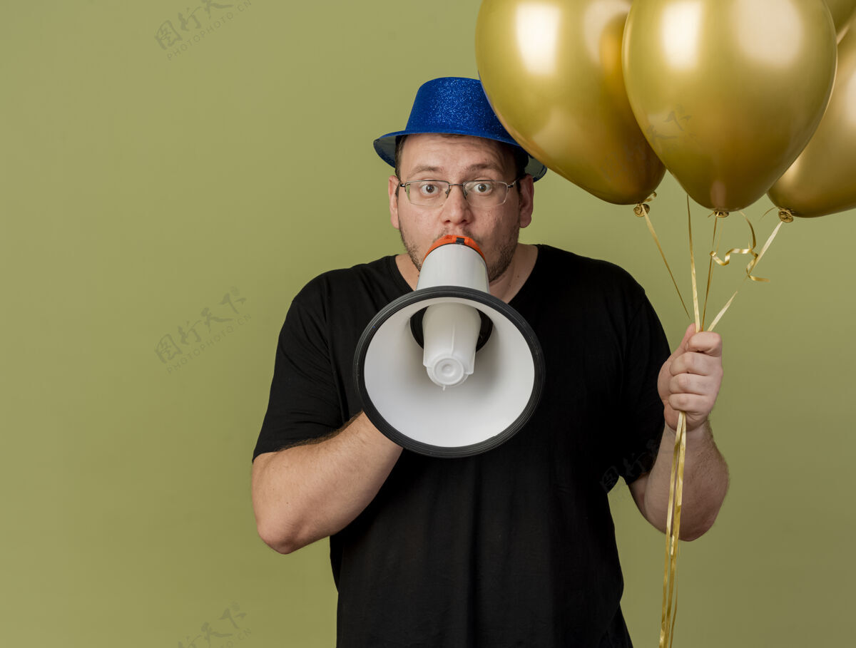 讲话一个戴着眼镜 戴着蓝色派对帽的成年男子手持氦气球 对着橄榄绿墙上孤立的扩音器讲话帽子斯拉夫气球