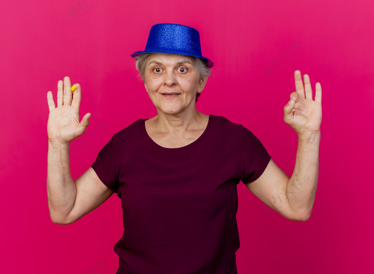脸高兴的老妇人戴着党的帽子站起来 举起的手拿着哨子和手势ok手的标志隔离在粉红色的墙上人穿上聚会