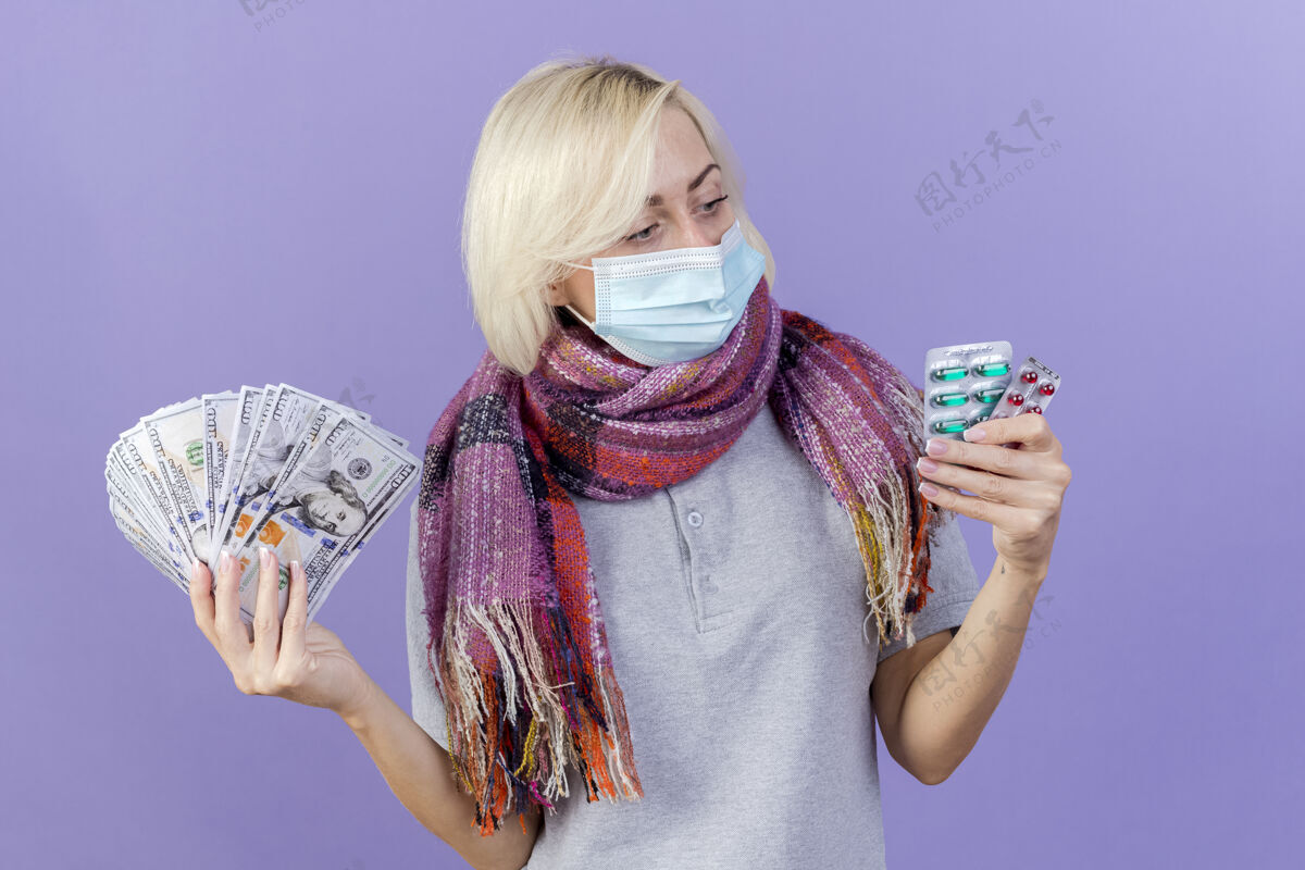 姿势戴着医用面罩和围巾的自信的金发病女人手里拿着钱 看着隔离在紫色墙上的几包药片脸金发面具