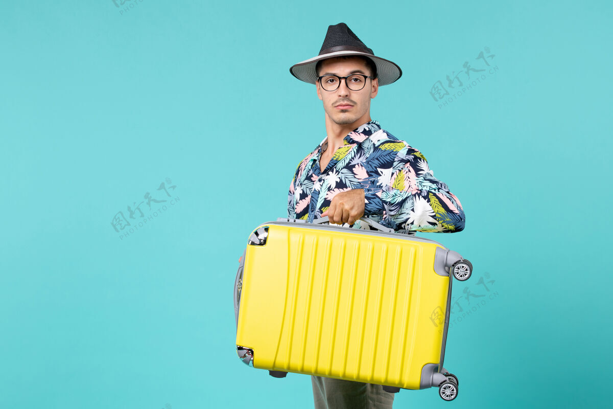 工作年轻的男性正准备旅行 拿着他的包在蓝色的墙上衬衫工人帽子