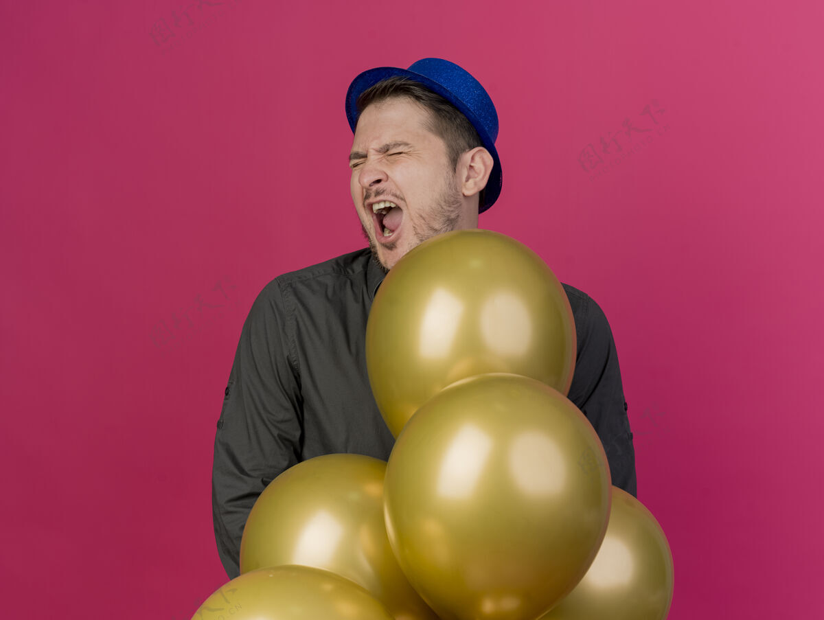 蓝色快乐地闭上眼睛 戴着蓝色帽子的年轻人站在粉红色的气球后面眼睛年轻气球