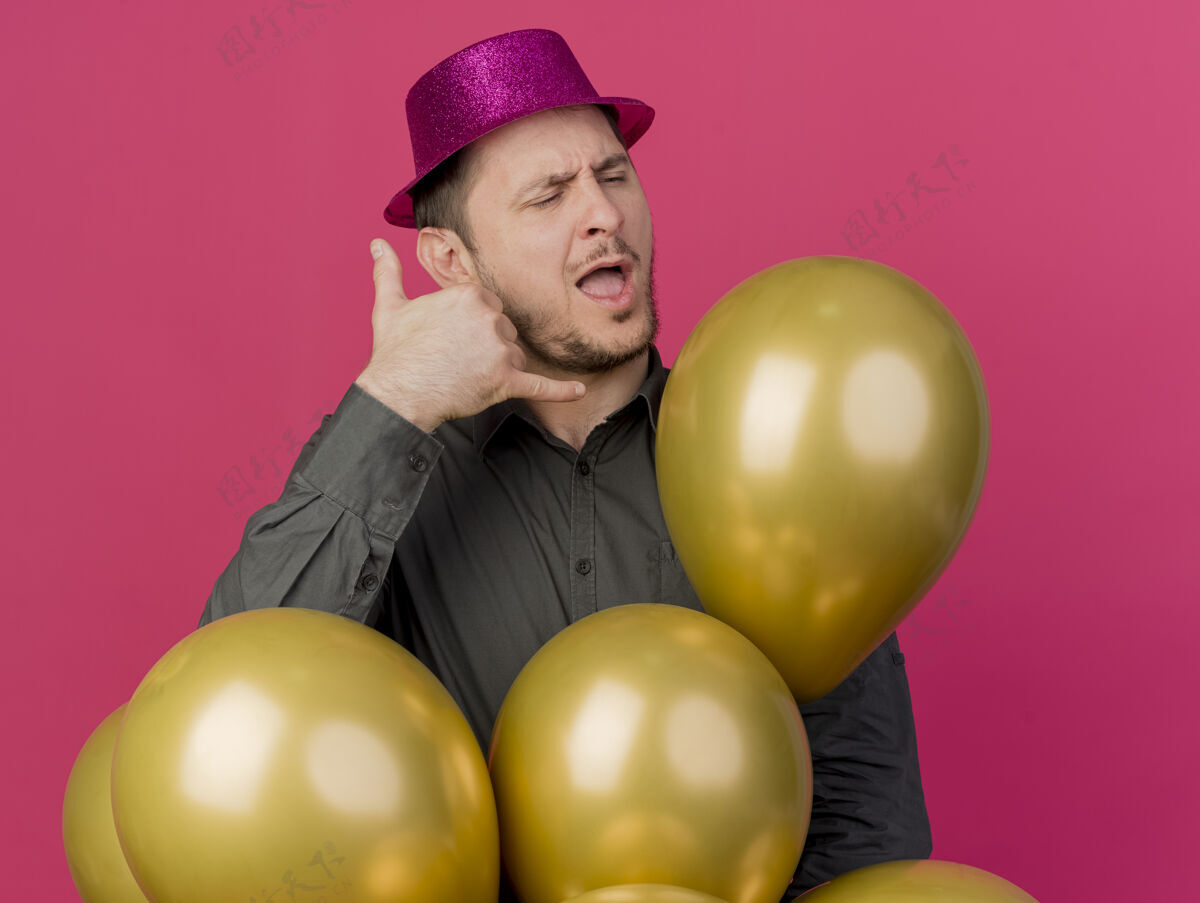 举行高兴的年轻人闭着眼睛戴着粉红色的帽子拿着气球打电话的手势孤立的粉红色穿家伙帽子