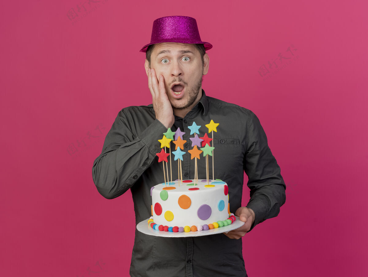 年轻害怕的年轻人戴着粉红色的帽子拿着蛋糕把手放在脸颊上孤立的粉红色帽子抱着脸颊