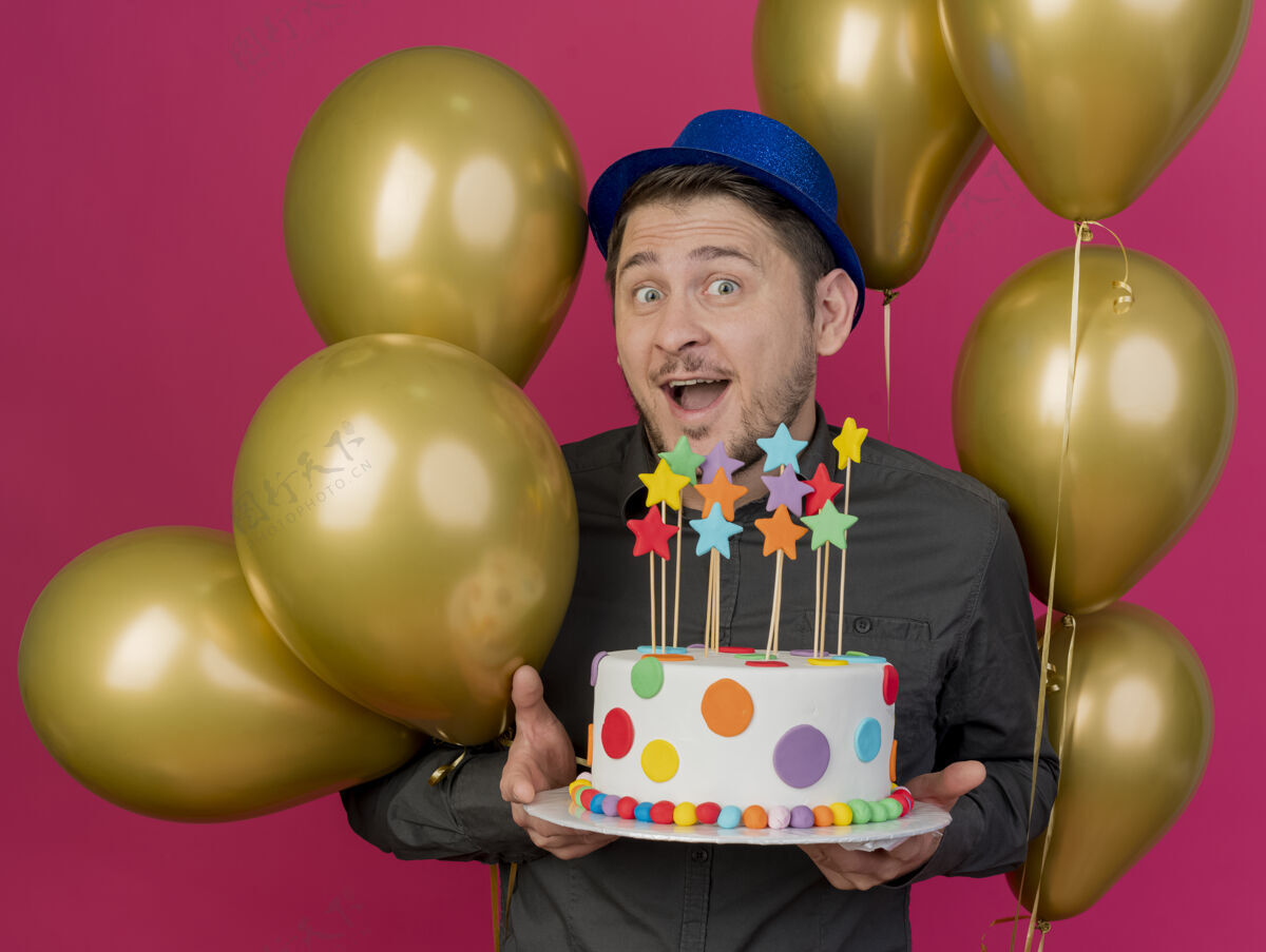 蛋糕快乐的年轻人戴着蓝色帽子站在气球中间 手里拿着粉红色的蛋糕高兴在其中年轻人