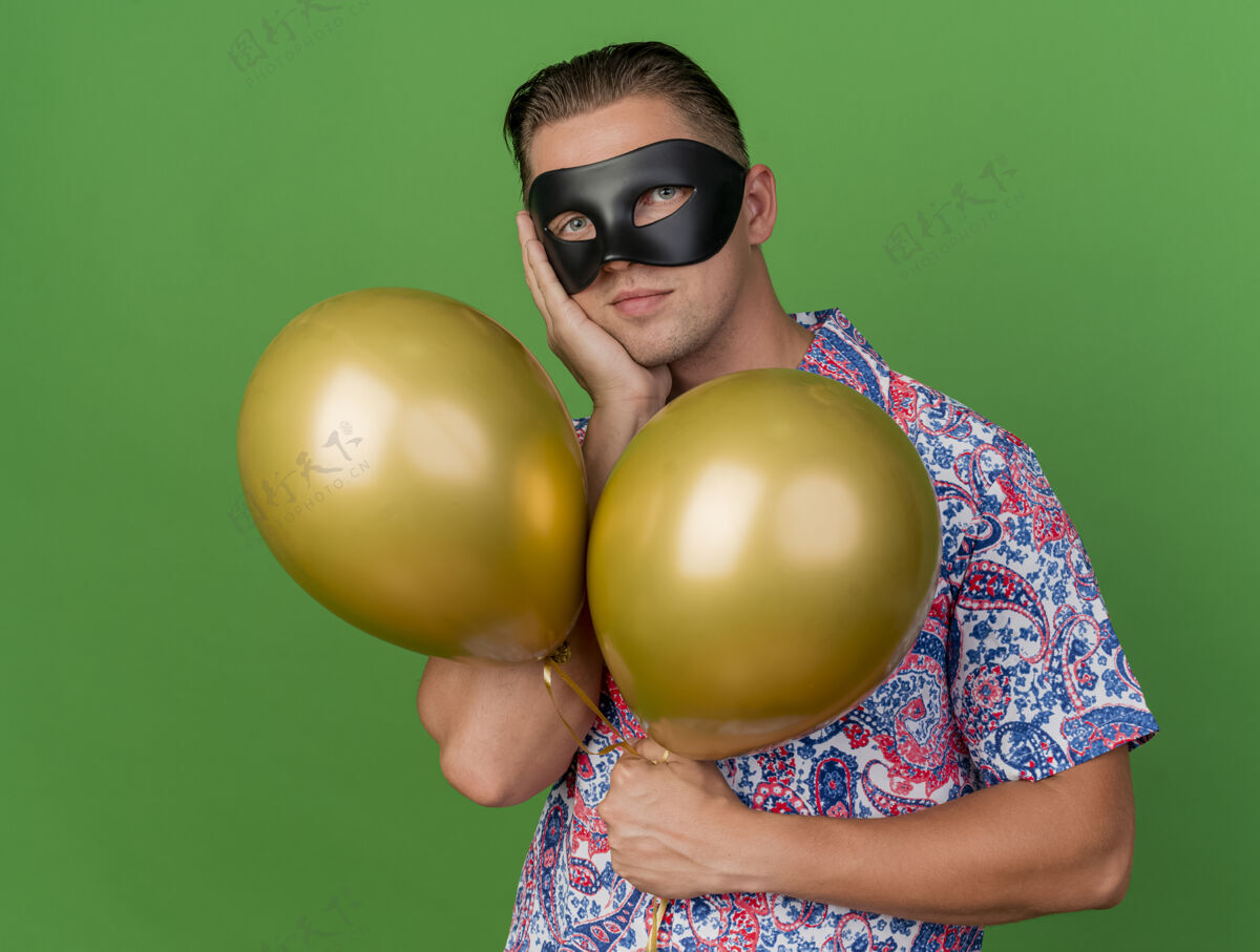 气球年轻人戴着化装眼罩拿着气球把手放在脸颊上隔离在绿色眼睛年轻化装舞会