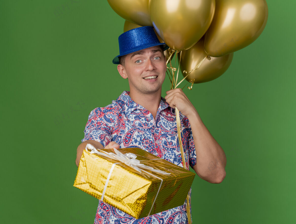 礼物面带微笑的年轻人戴着蓝色的帽子拿着气球和礼品盒孤立地站在绿色戴着年轻人拿着