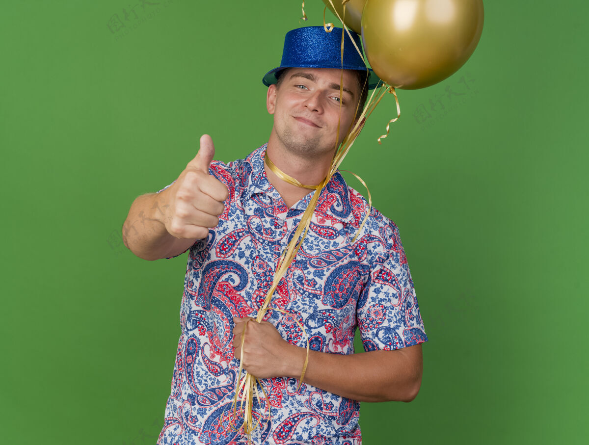 表演高兴的年轻人戴着蓝色的帽子 脖子上绑着气球 在绿色的草坪上竖起大拇指请小伙子拇指