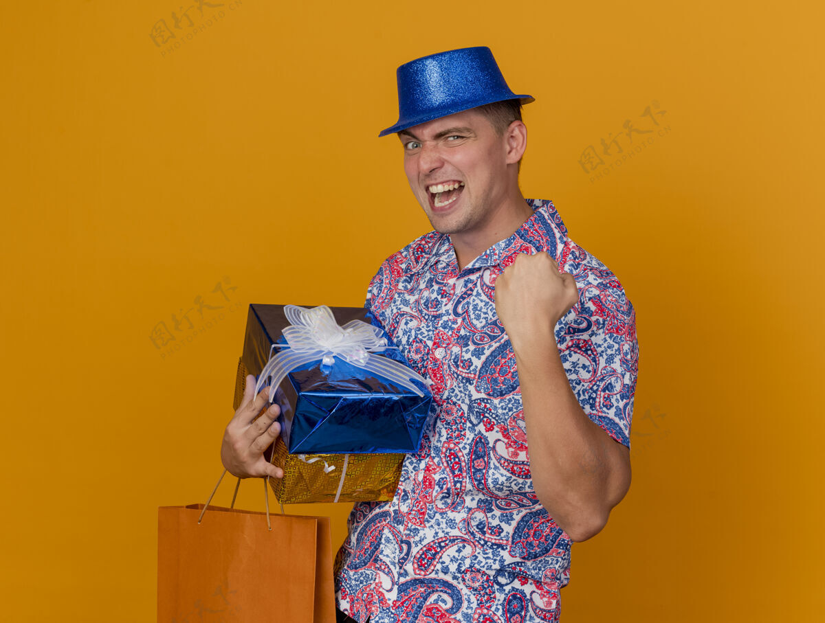 是的快乐的年轻人戴着蓝色的帽子 拿着礼品盒和袋子 在橙色上显示“是”的手势包小伙子穿