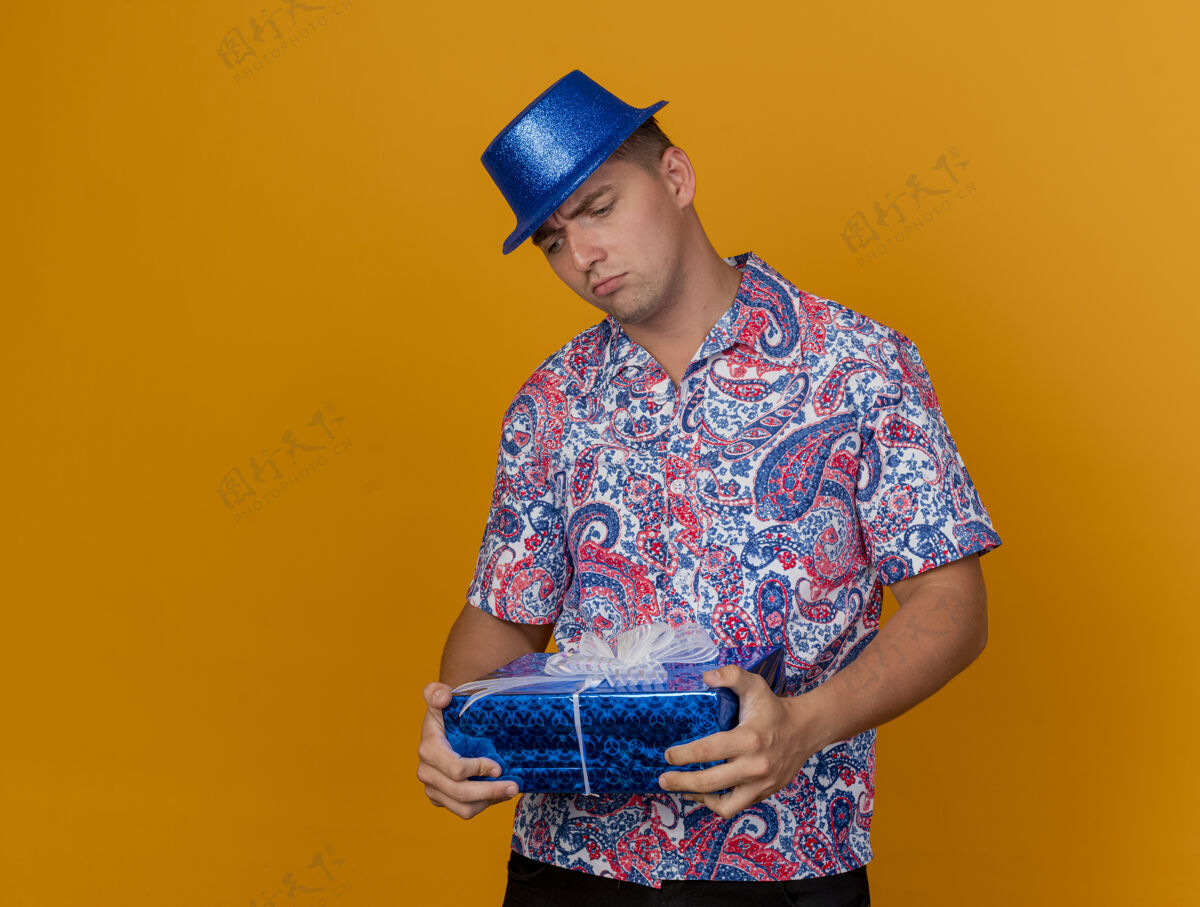 橙色一个体贴的年轻人戴着蓝色的帽子 拿着礼物盒 看着孤立的橙色思考小伙子盒子