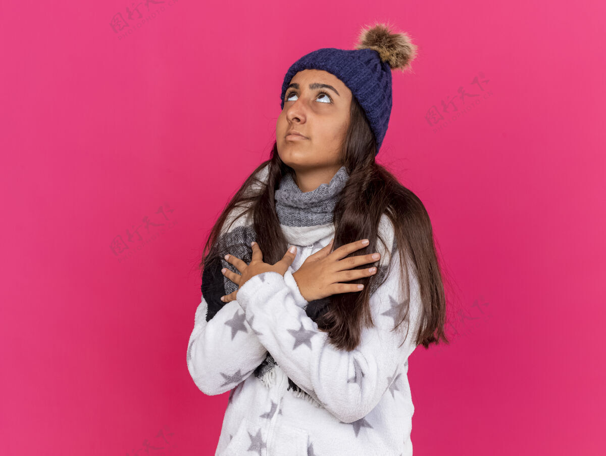 穿年轻生病的女孩戴着冬天的帽子 戴着围巾 双手放在胸前 隔离在粉红色的衣服上粉色围巾帽子