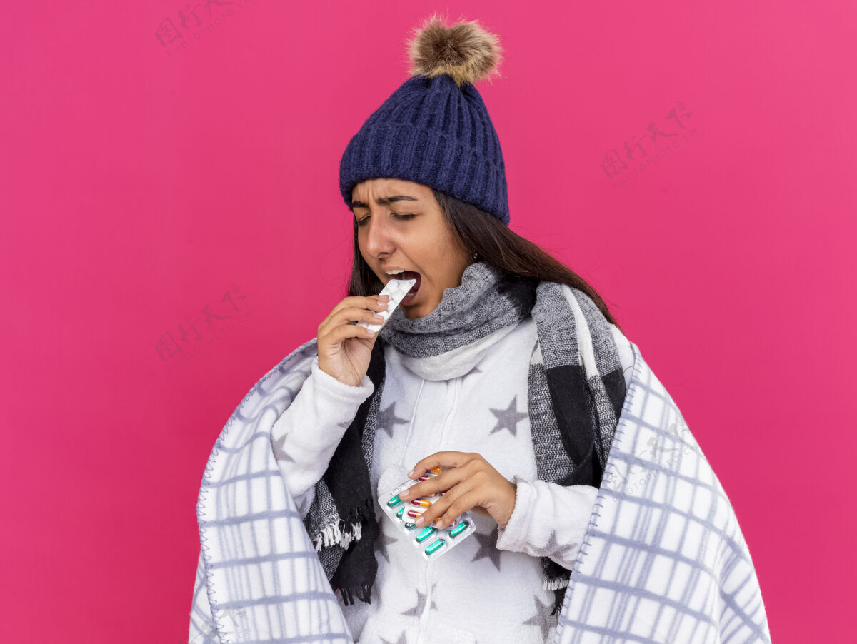 药片年轻的病女孩闭着眼睛 戴着冬天的帽子 围巾裹着格子布 拿着药片 把药片放进嘴里 隔离在粉红色的衣服上眼睛封闭持有
