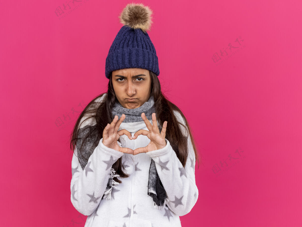穿悲伤的生病的年轻女孩戴着冬天的帽子 戴着围巾 在粉红色的脸上露出心形手势疾病帽子