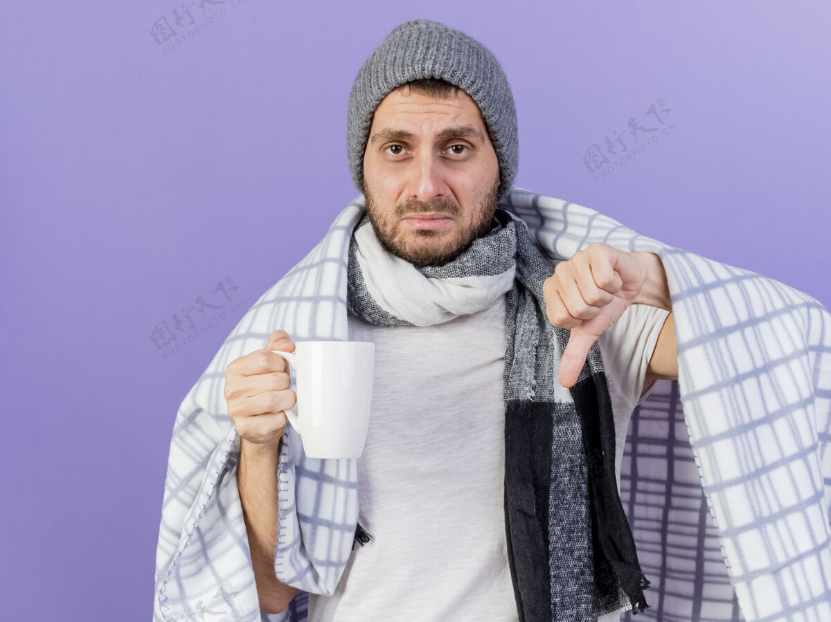 杯子悲伤的年轻病夫戴着冬天的帽子 戴着围巾 端着一杯茶 大拇指朝下 孤零零地站在紫色上茶帽子拇指