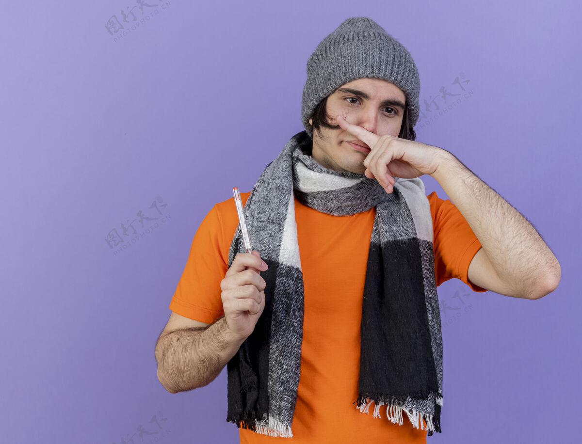 围巾年轻的病夫戴着冬天的帽子 戴着围巾 拿着温度计 用手指擦鼻子 把紫色隔离疾病壁板体温计