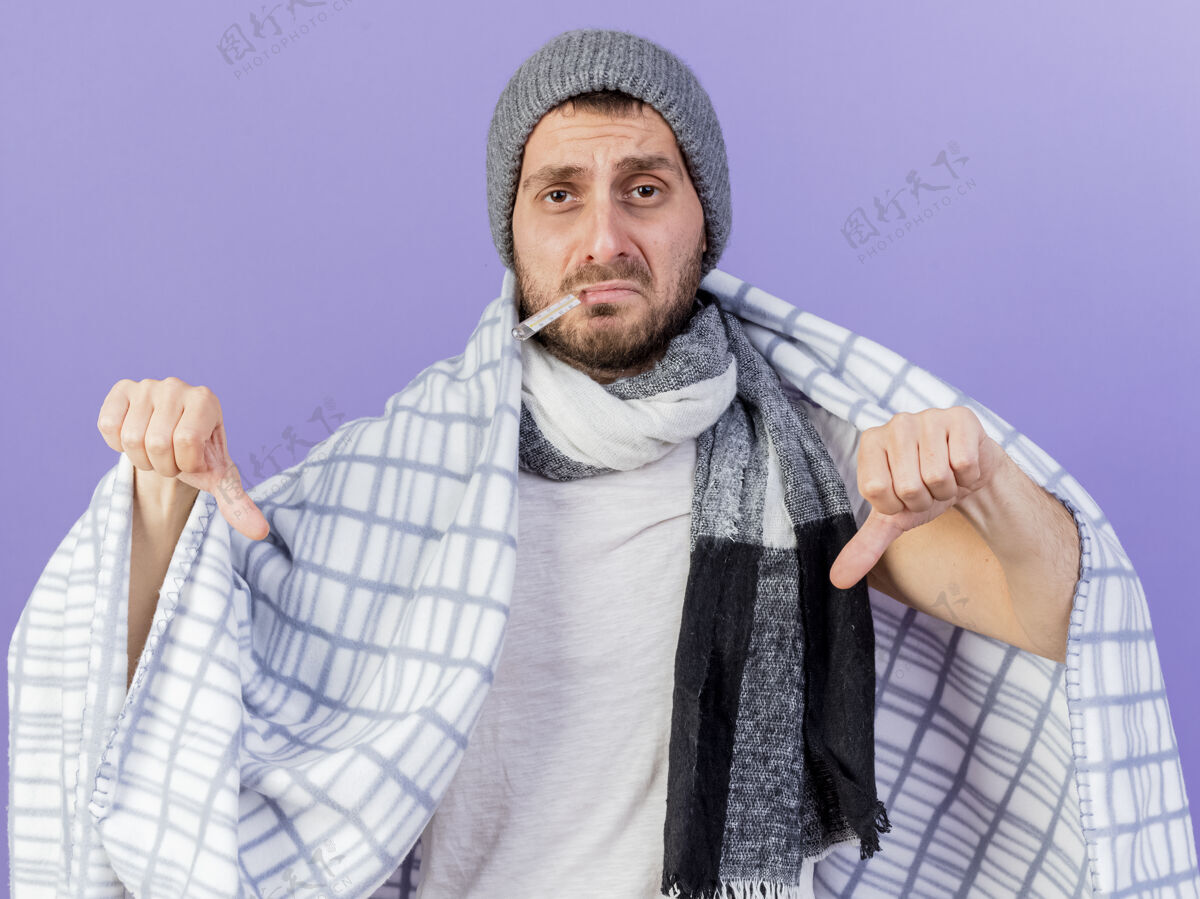 格子悲伤的年轻病人戴着冬天的帽子 戴着围巾 把体温计放进嘴里 用格子布包着 大拇指朝下 孤立地站在紫色地上帽子展示包装