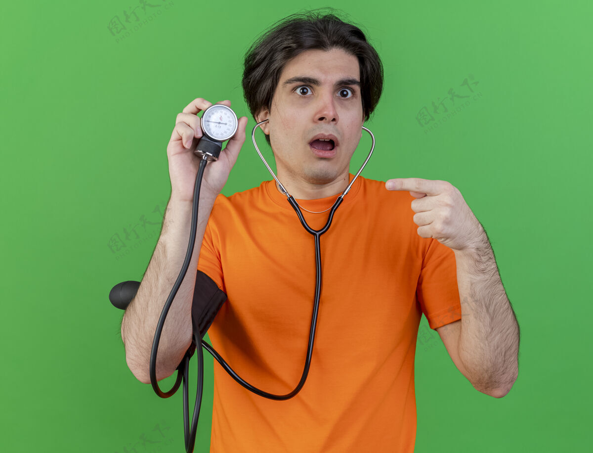 惊喜惊讶的年轻人戴着听诊器拿着血压计指着绿色隔离年轻戴男子