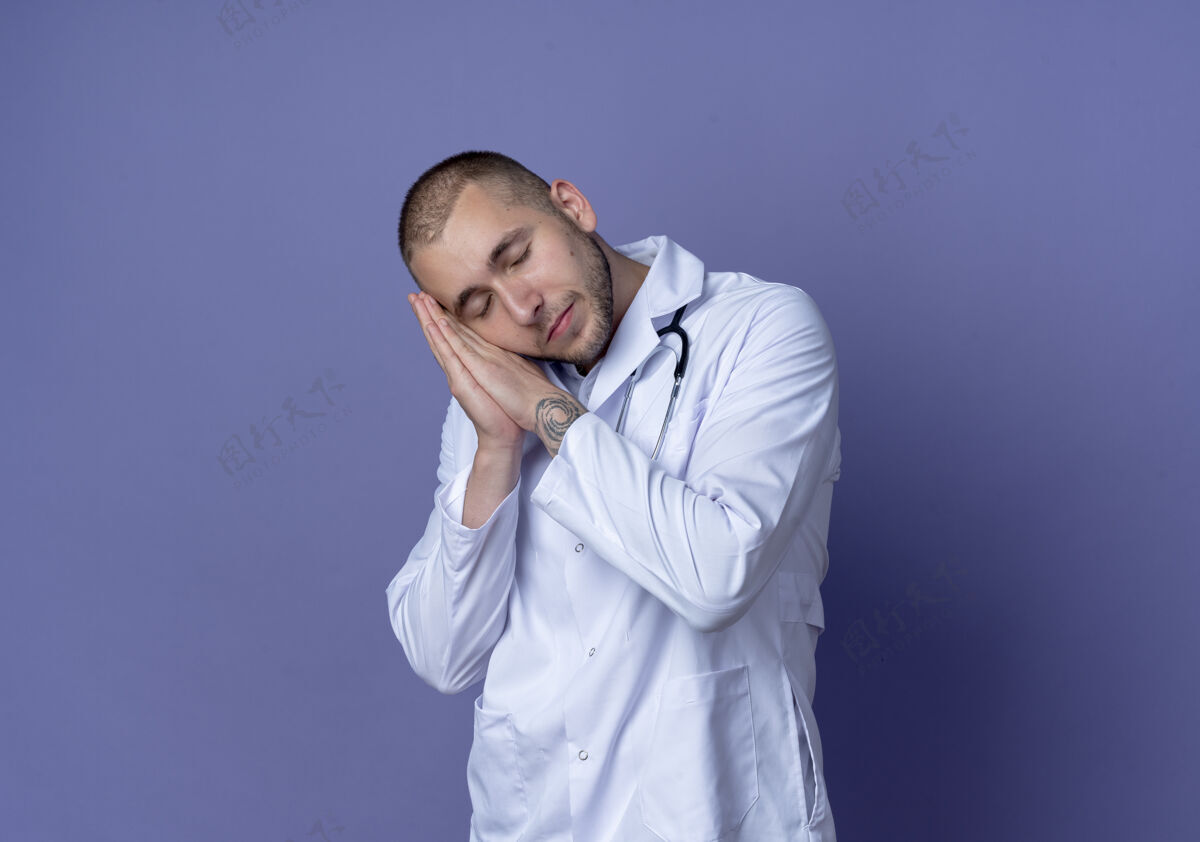 穿着昏昏欲睡的年轻男医生 穿着医用长袍 戴着听诊器 闭着眼睛在紫色的墙上做着睡觉的姿势医生长袍表情