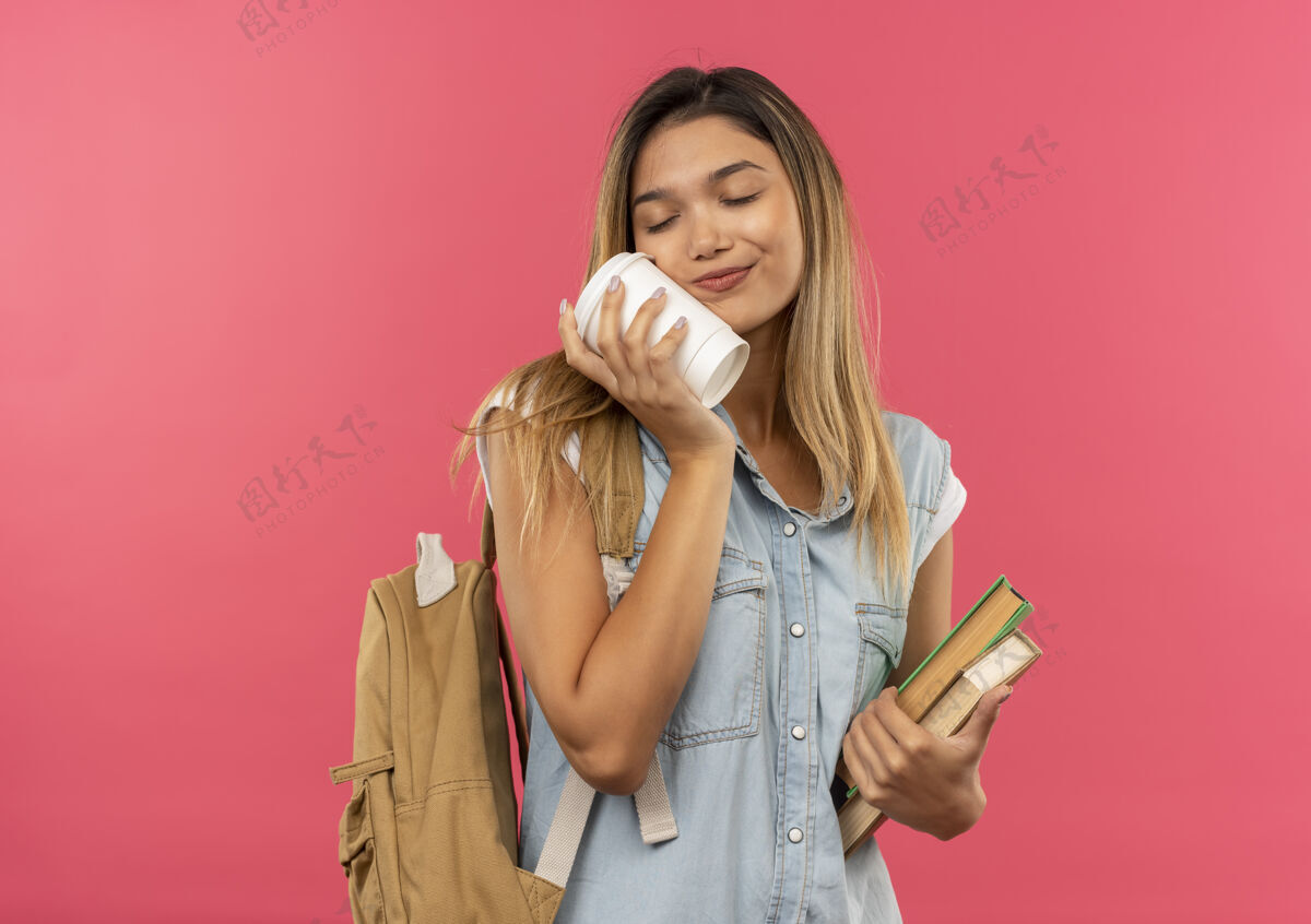 女孩高兴的年轻漂亮的女学生戴着背袋拿着书 用闭着眼睛的塑料咖啡杯摸着脸 隔离在粉红色的墙上闭着眼睛年轻眼睛人