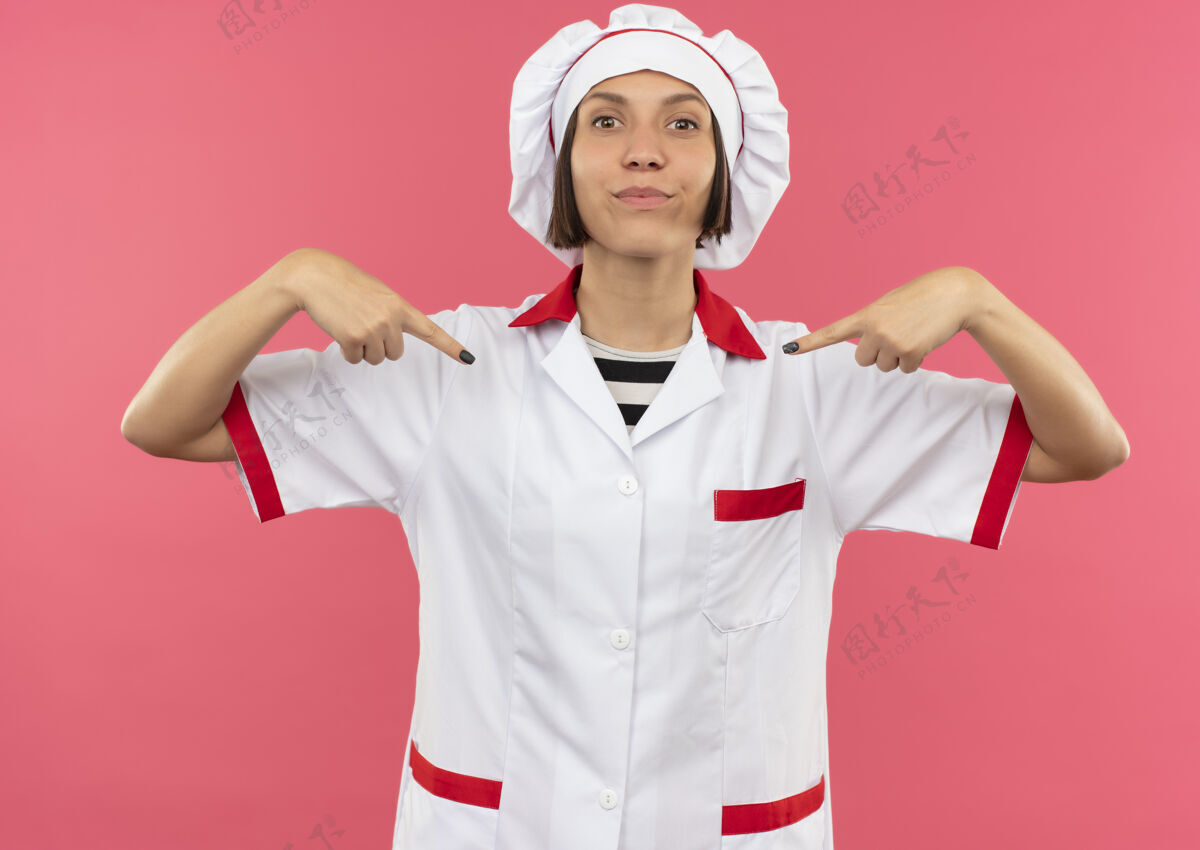 她自己身着厨师制服的年轻女厨师高兴地指着隔离在粉红色墙上的自己人衣服厨师