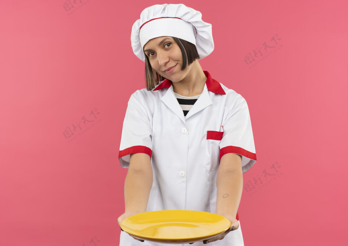 人身着厨师制服的年轻女厨师高兴地把盘子朝前伸出 孤立地放在粉红色的墙上表情衣服烹饪