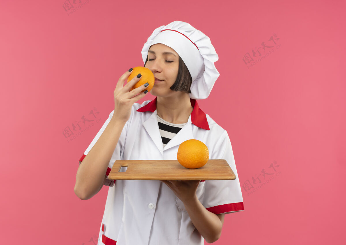 人身着厨师制服的年轻女厨师手拿着插着橘子的砧板 闭着眼睛在粉红色的墙上嗅着橘子味表情请公民