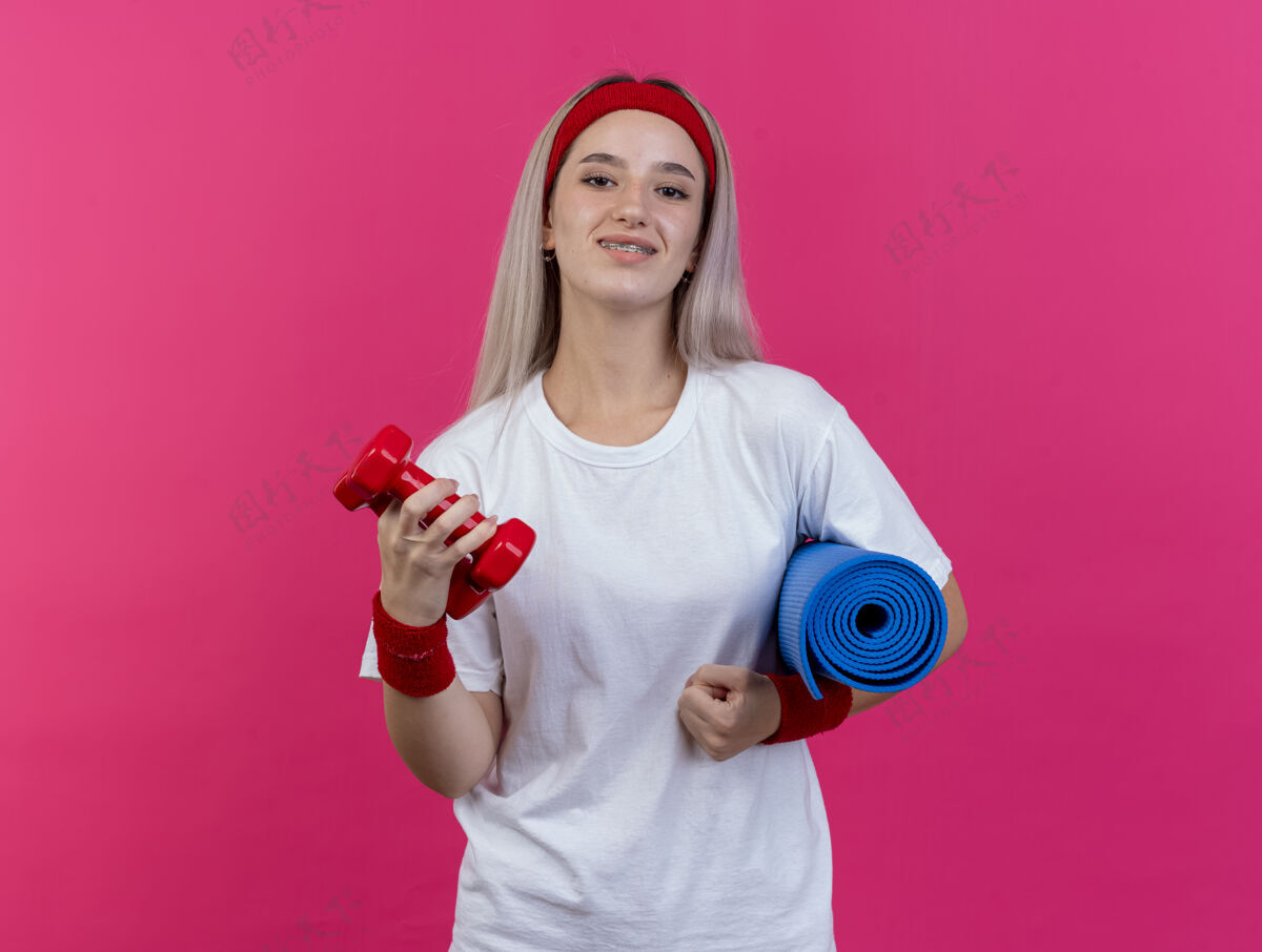 头带面带微笑的年轻运动女性戴着背带 戴着头带和腕带 拿着运动垫和哑铃隔离在粉红色的墙上运动微笑女人
