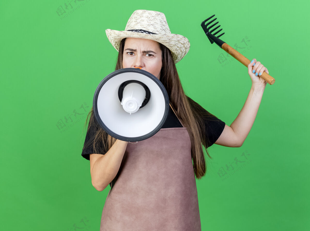 绿色穿着制服的严格漂亮的园丁女孩戴着园艺帽在喇叭上讲话 在绿色上举起耙子严格说话喇叭