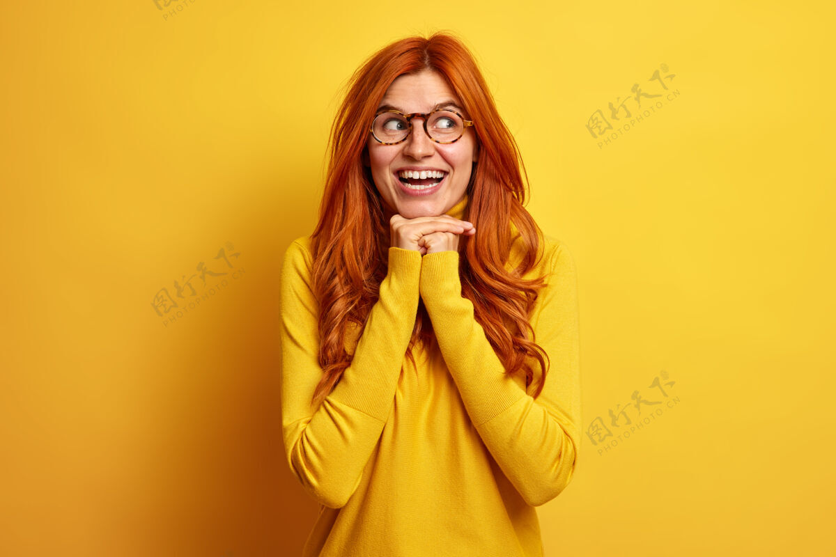 欢呼漂亮的红发欧洲女人的画像笑容灿烂双手放在下巴下高兴地看着一旁穿着休闲套头衫表示幸福休闲年轻高兴