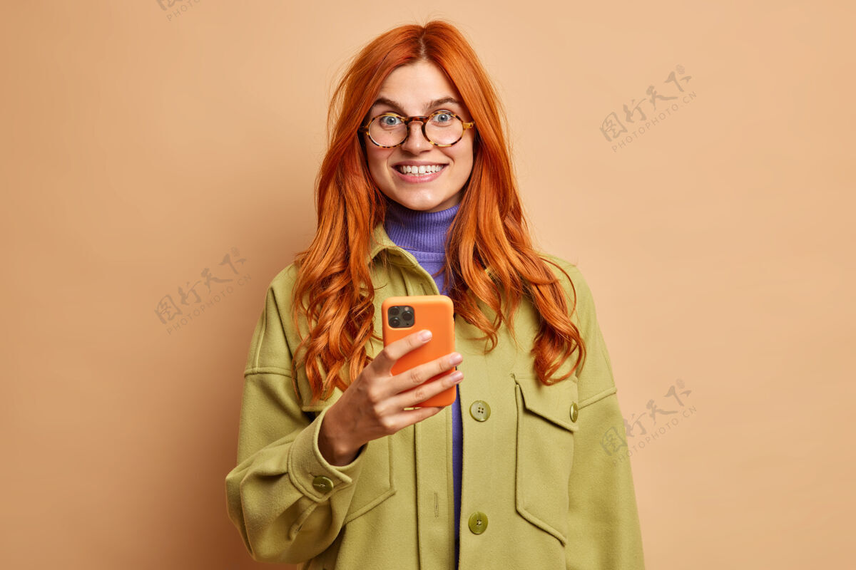 白种人移动服务和在线通讯概念穿着时髦的秋季夹克 高兴的漂亮红发女青年手捧着现代手机 咧嘴笑着眼镜年轻女士