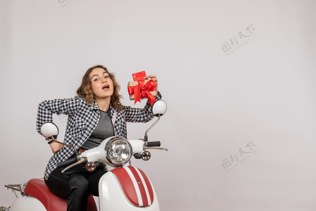 音乐前视图可爱的年轻女子骑着轻便摩托车拿着礼物和卡片在灰色的墙上举行前面快乐