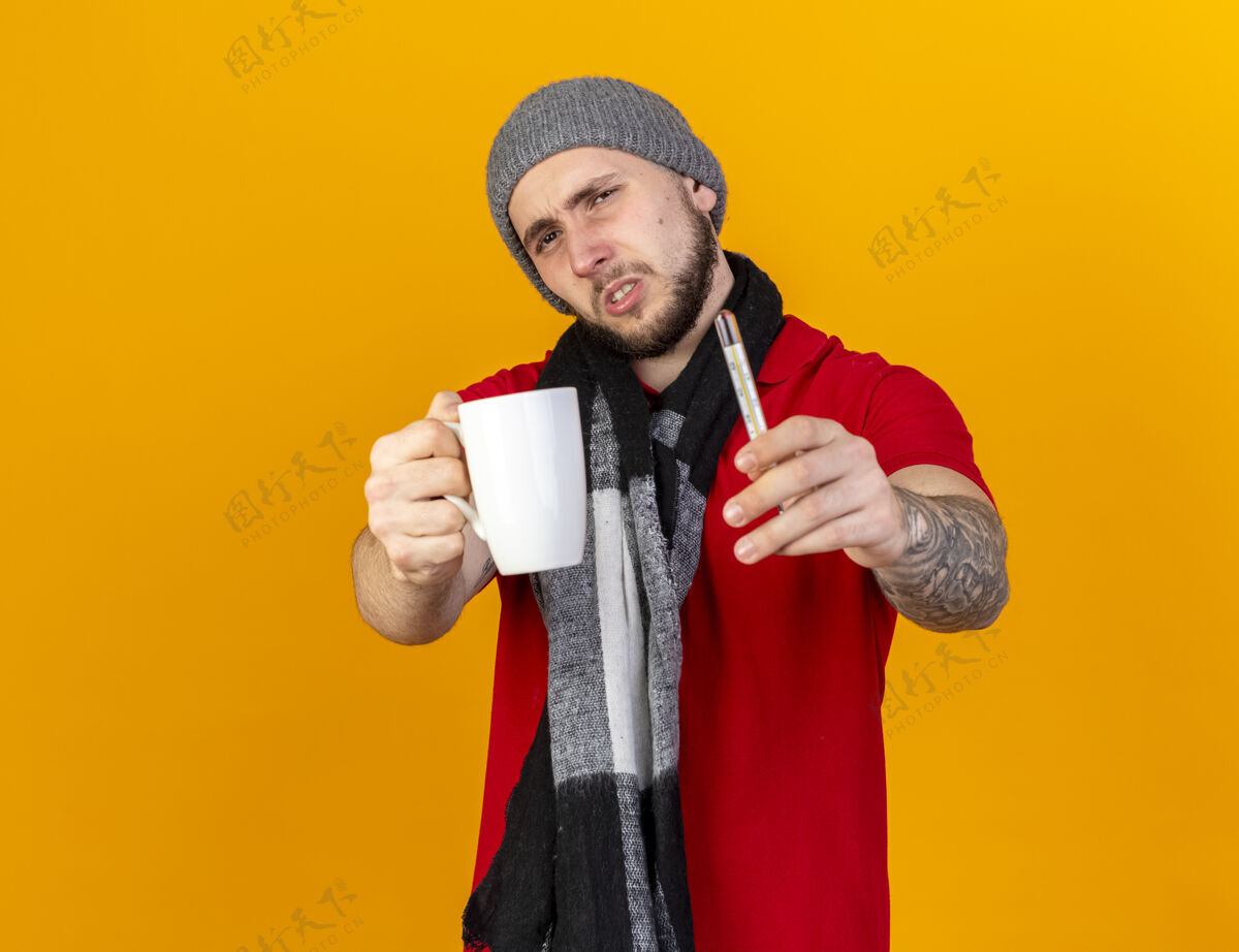 帽子年轻的白种人 戴着冬天的帽子 戴着围巾 拿着杯子和温度计 隔离在橙色的墙上 留着复印空间疾病不愉快体温计