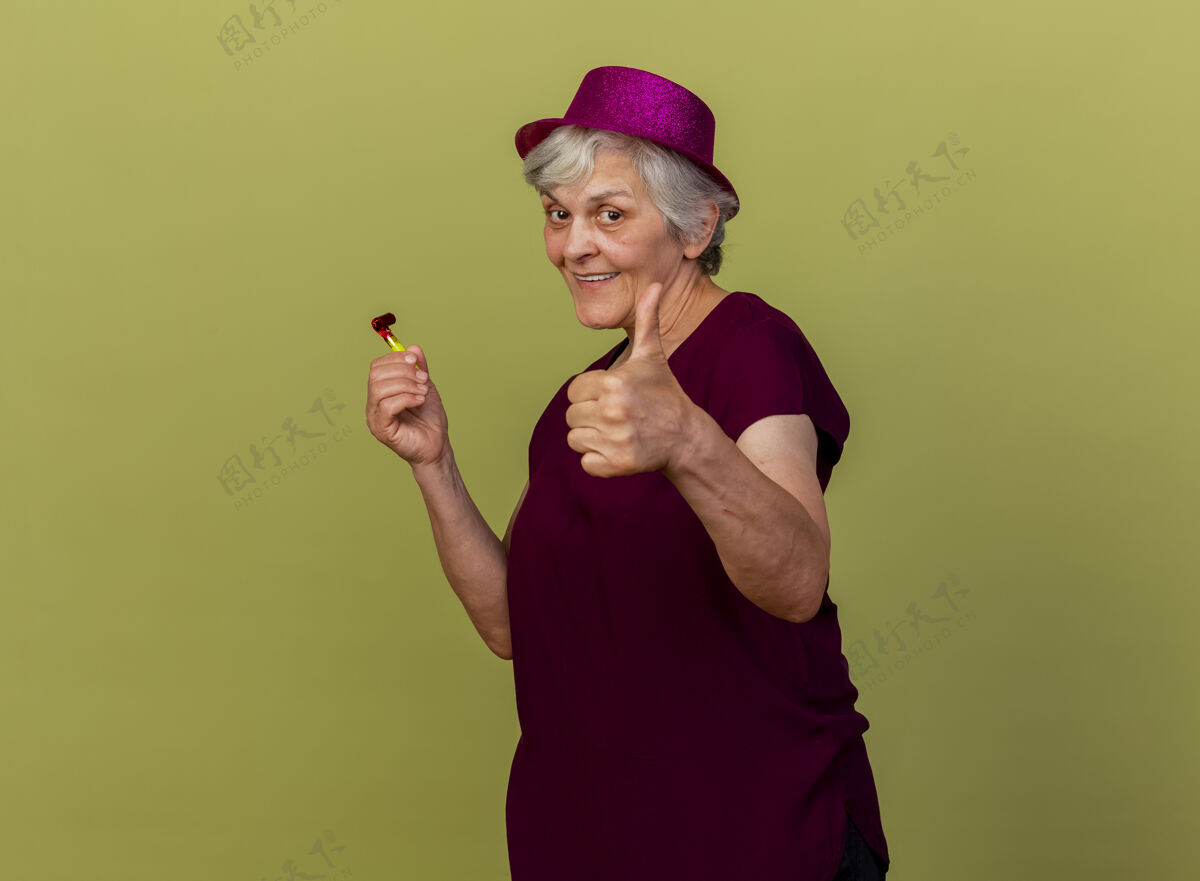 戴面带微笑的戴着派对帽的老妇人竖起大拇指 举着隔离在橄榄绿墙上的口哨 还有复印空间女人帽子口哨