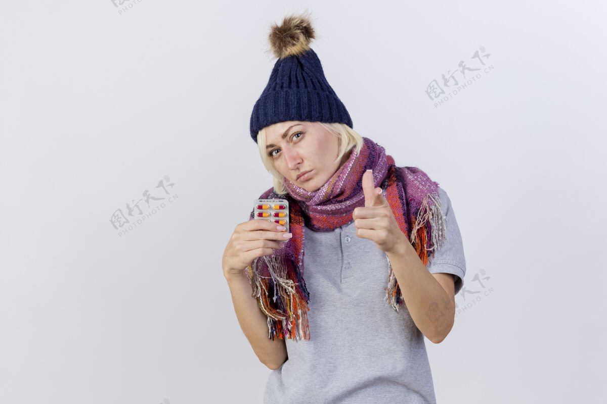 穿自信的年轻金发病斯拉夫妇女戴着冬天的帽子和围巾 持有一包药片和点隔离在白色墙上的复制空间围巾女人年轻