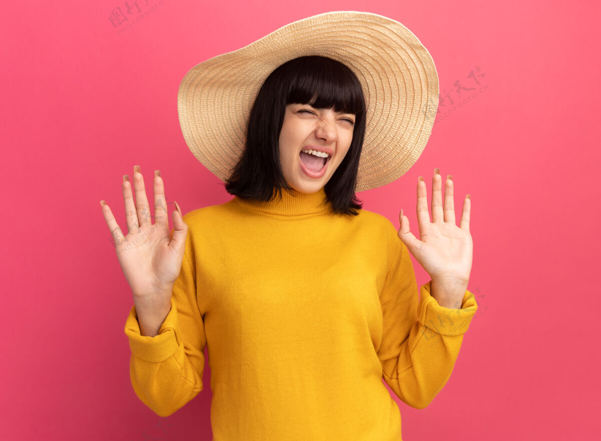 帽子恼怒的年轻黑发高加索女孩戴着沙滩帽站在粉红色上举手黑发站着年轻人