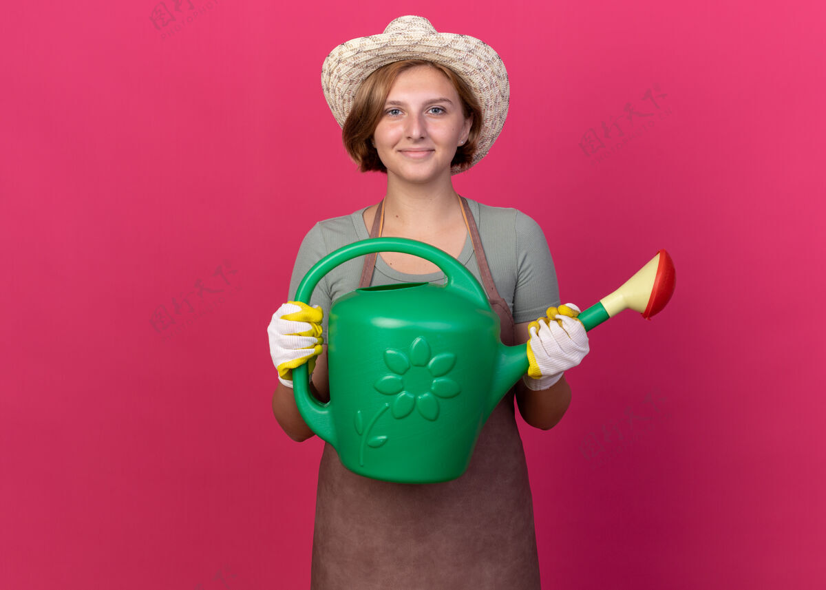 园艺高兴的年轻斯拉夫女园丁戴着园艺帽和手套拿着粉红色的浇水罐花园女的手套