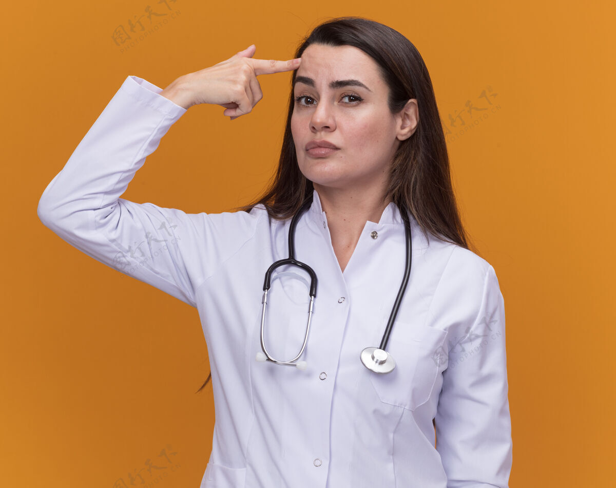 长袍自信的年轻女医生穿着带听诊器的医用长袍 把手指放在橙色的额头上听诊器年轻医学