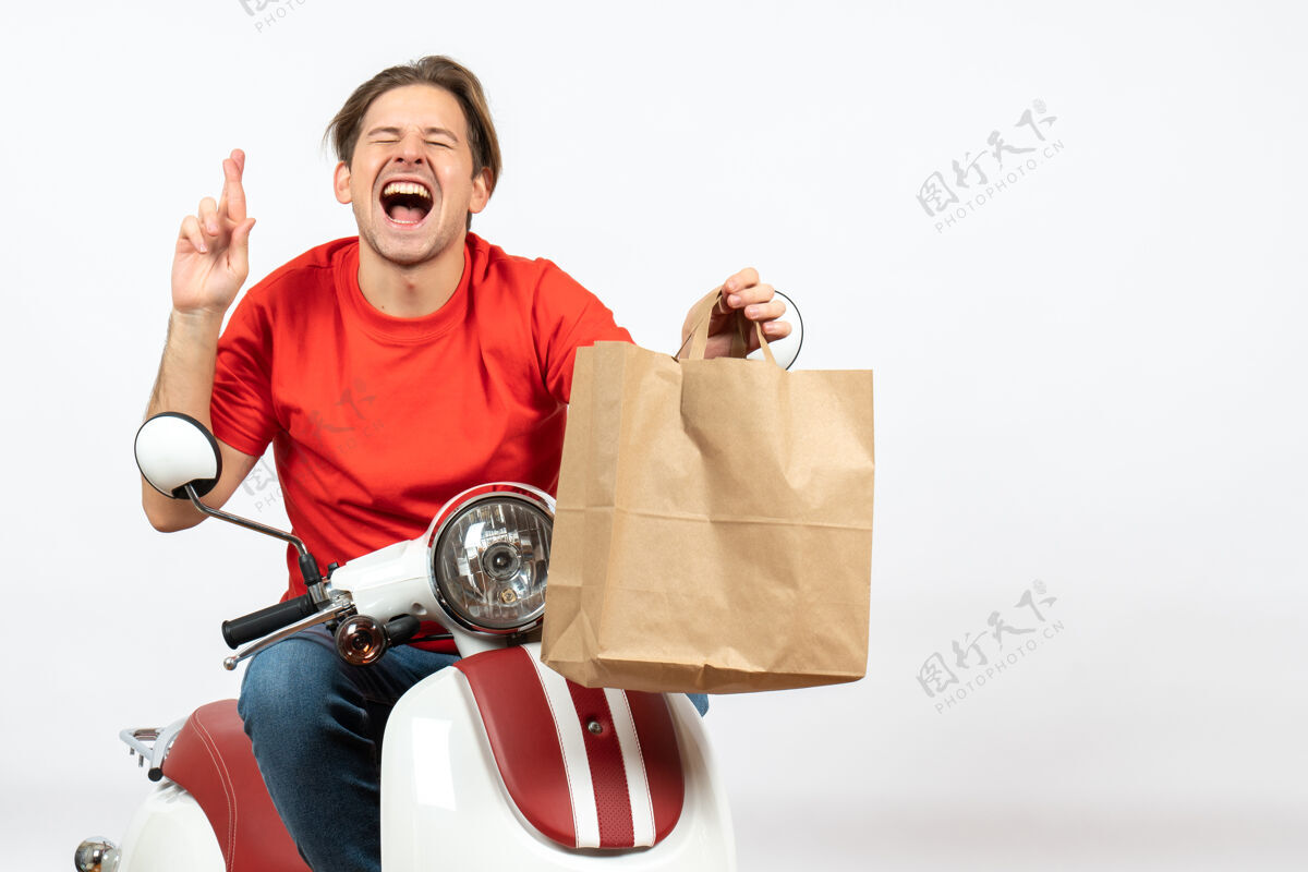 男人年轻的充满希望的情绪快递员穿着红色制服坐在踏板车上给白色墙上的纸袋人情感生意