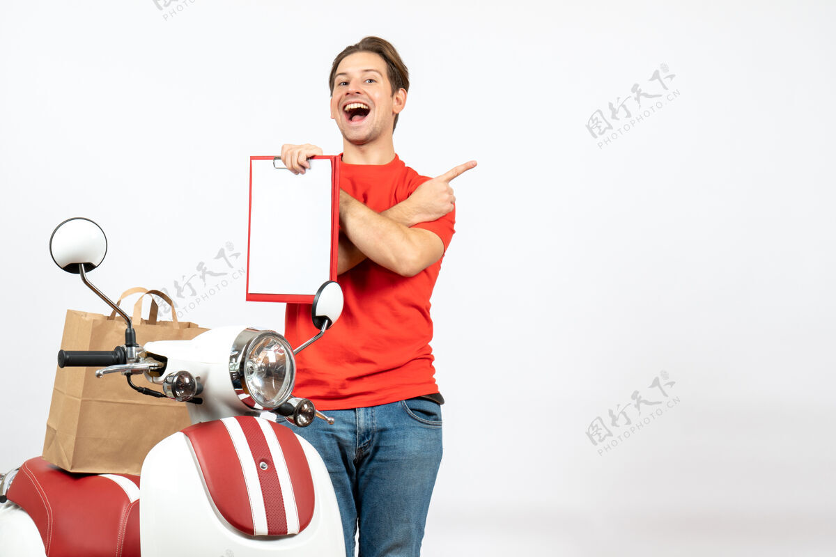 站立俯视图中 身着红色制服 面带微笑的年轻送货员站在滑板车旁 展示文件 并指向白墙上的背面快乐展示文件