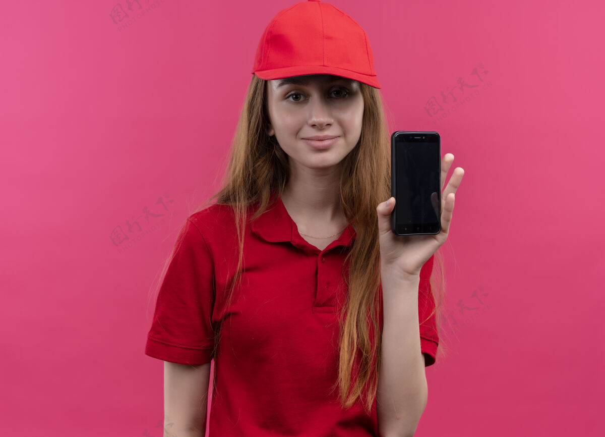 送货一个穿着红色制服 拿着手机的年轻送货女孩高兴地站在孤立的粉色空间里女孩年轻制服
