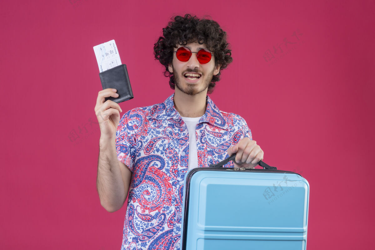 英俊快乐的年轻帅气的卷发旅行者戴着太阳镜拿着钱包和带手提箱的机票在隔离的粉红色空间和复印空间男人快乐机票