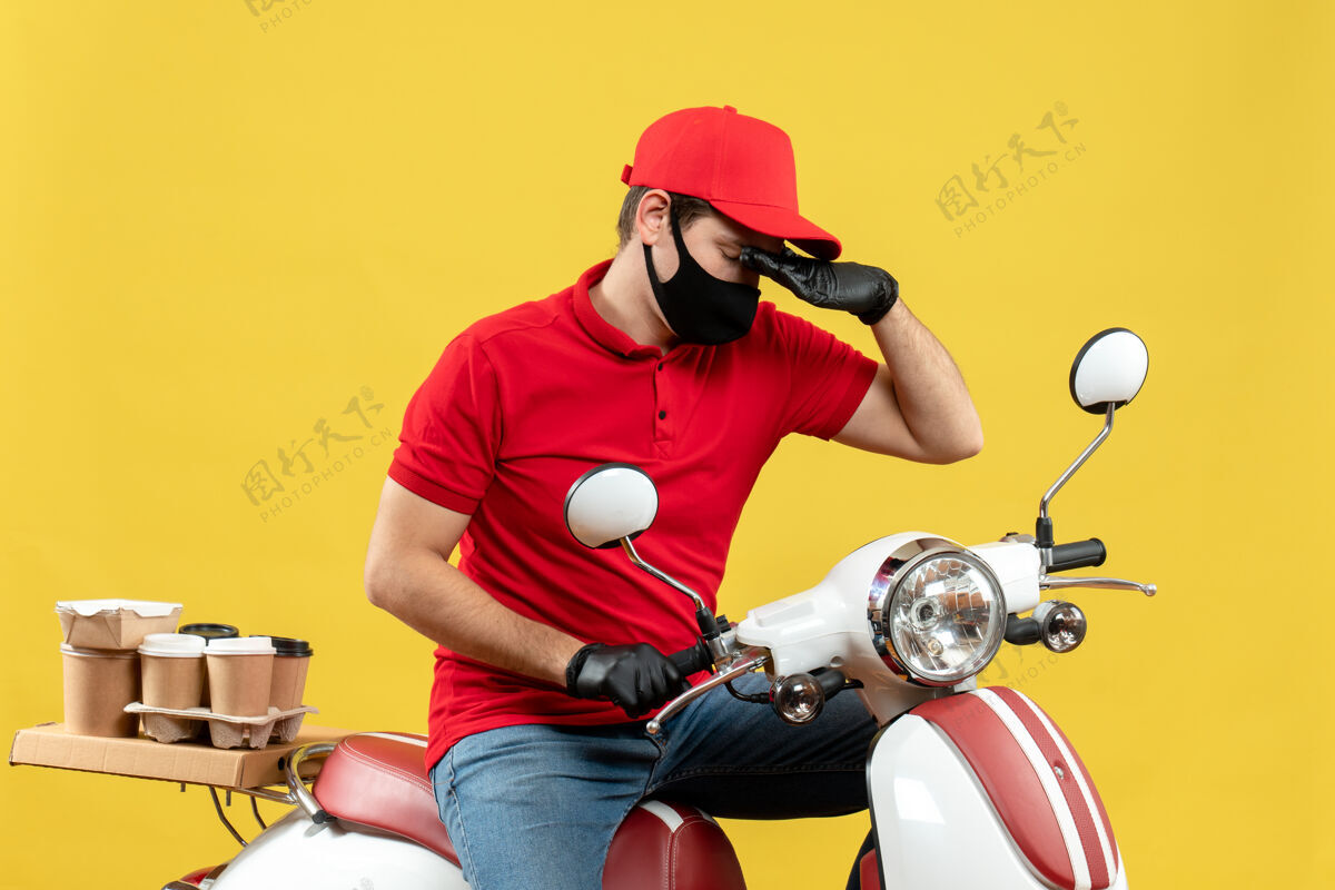 订单俯视图：穿着红色上衣 戴着帽子手套 戴着医用面罩 坐在黄色墙上的滑板车上传递命令的年轻人滑板车年轻人手套