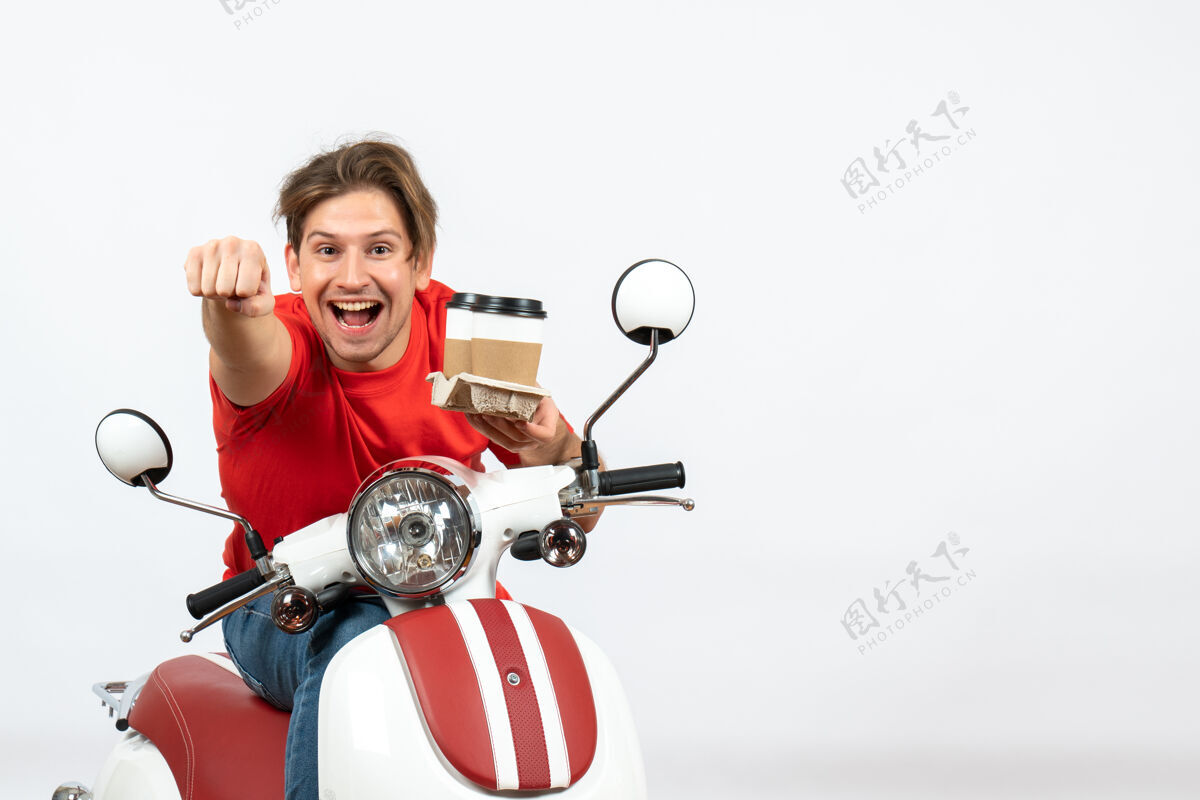 摩托车穿着红色制服的年轻快乐的快递员坐在踏板车上 在黄色的墙上拿着订单年轻人举行男人