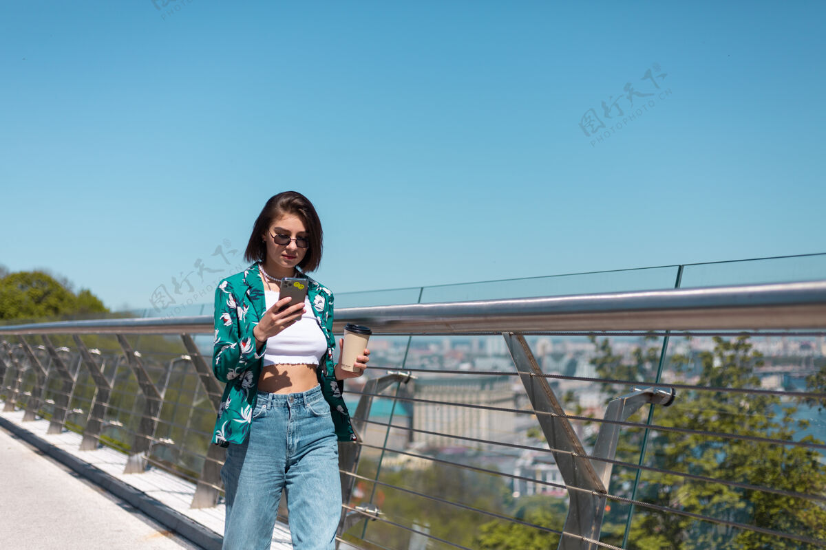 复制空间阳光明媚的日子里 穿着休闲绿衬衫和牛仔裤的女人在桥上散步 手机屏幕上放着一杯咖啡墙电话女士